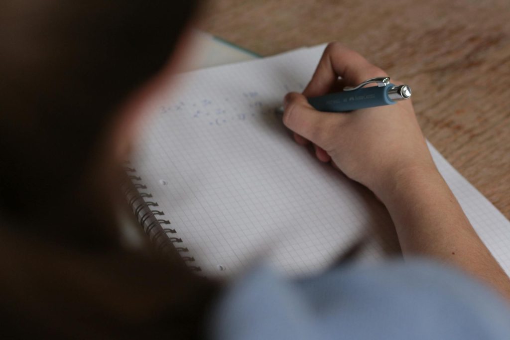 Una mano escribiendo fórmulas matemáticas en un cuaderno de cuadrícula con un bolígrafo azul.