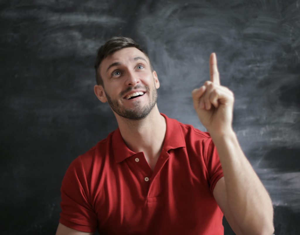 Un hombre sonriente con polo rojo señala con el dedo hacia arriba, simulando una idea o un momento de inspiración frente a un fondo oscuro.